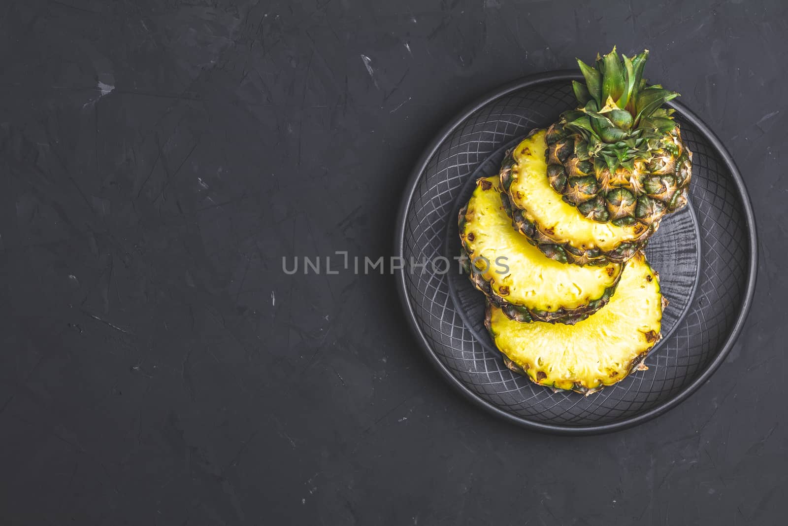 Sliced pineapple in ceramic plate on black stone concrete textured surface background. Top view with copy space for your text.