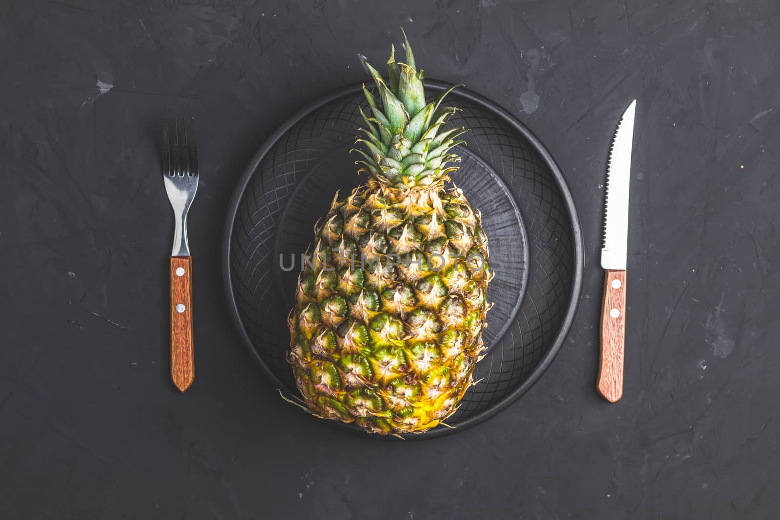 Pineapple in ceramic plate and set of cutlery knife, fork  on black stone concrete textured surface background. Top view with copy space for your text.