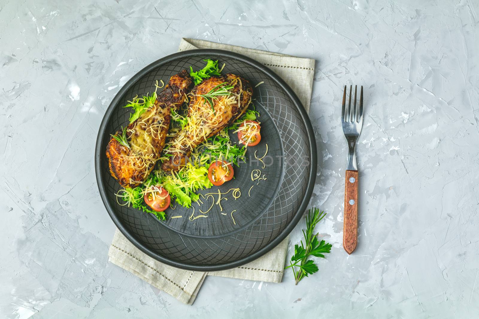 Baked chicken drumstick in a black ceramic plate with orange and rosemary, light gray stone concrete surface, top view, copy space.