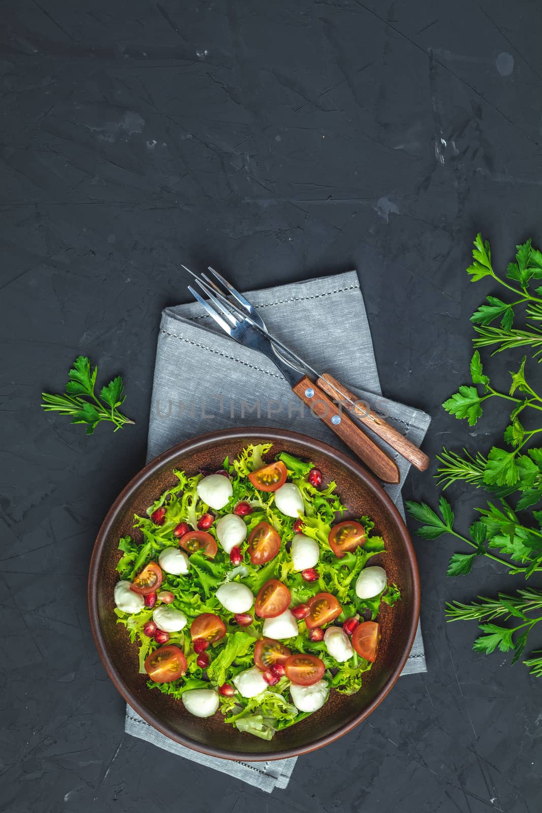 Fresh Cherry Tomato, Mozzarella salad with green lettuce mix served on a brown ceramic plate, healthy food, black stone concrete surface, top view, copy space.