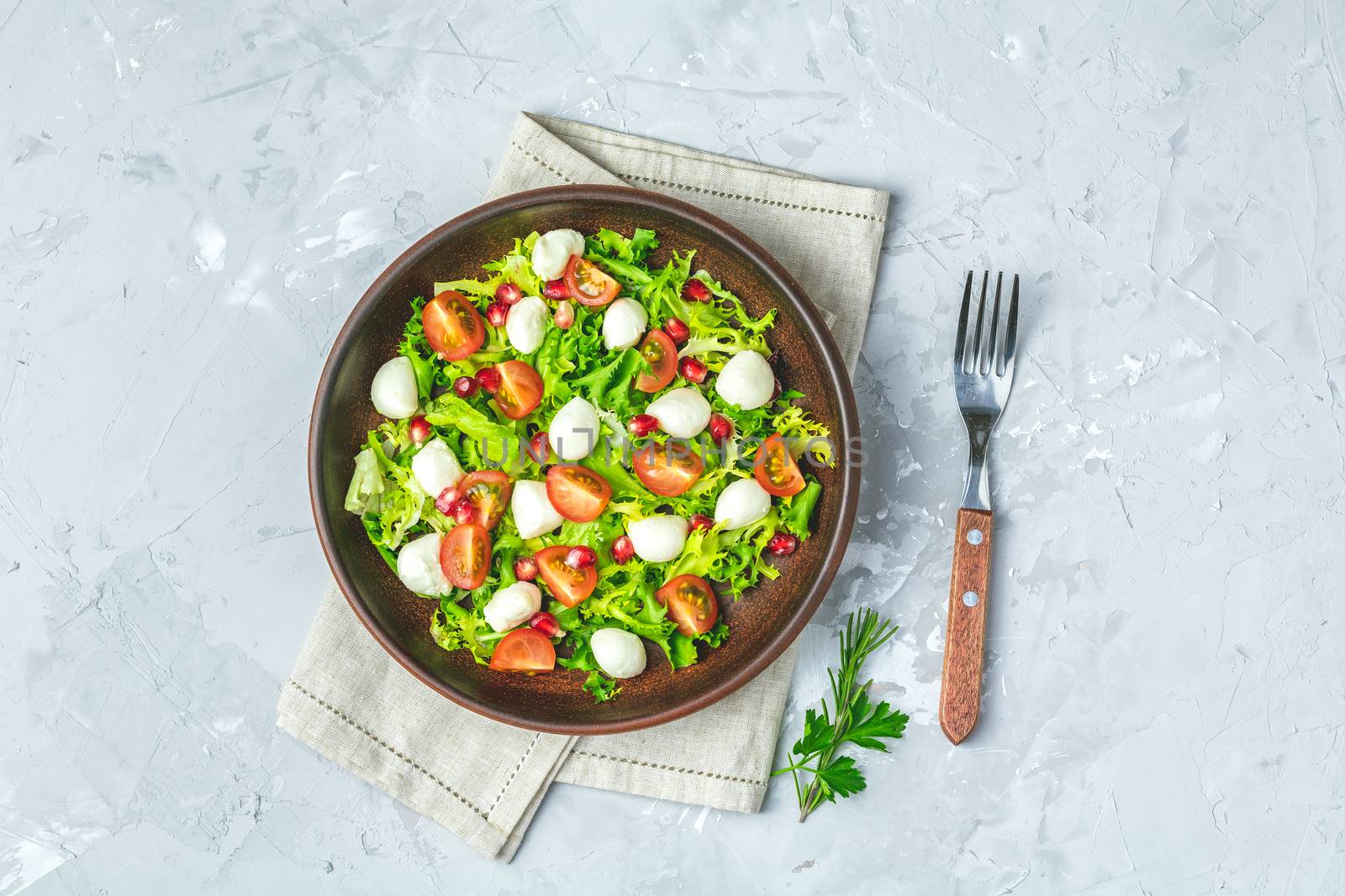 Fresh Cherry Tomato, Mozzarella salad with green lettuce mix served on a brown ceramic plate, healthy food, light gray stone concrete surface, top view, copy space.