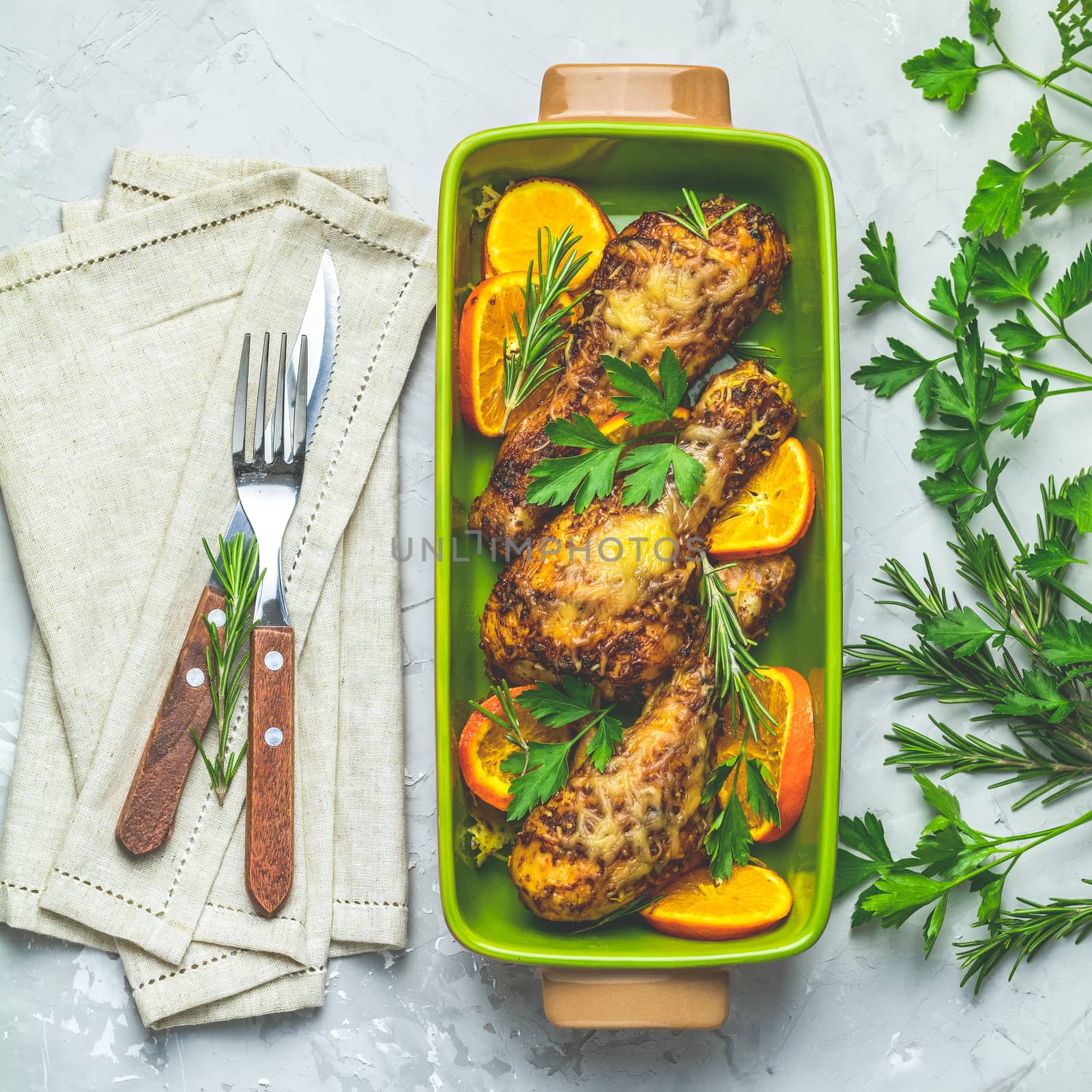 Baked chicken drumstick in a green dish with orange and rosemary, light gray stone concrete surface, top view, copy space.