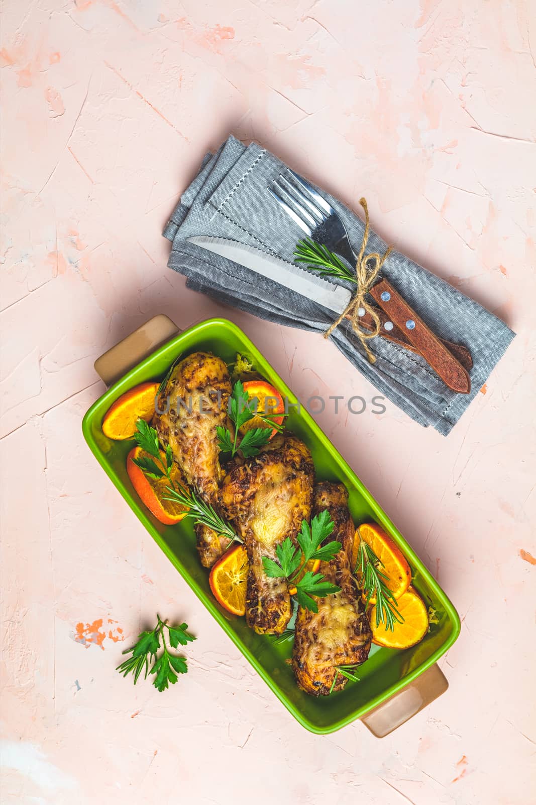 Baked chicken drumstick in a green dish with orange and rosemary, light pink living coral stone concrete surface, top view, copy space.