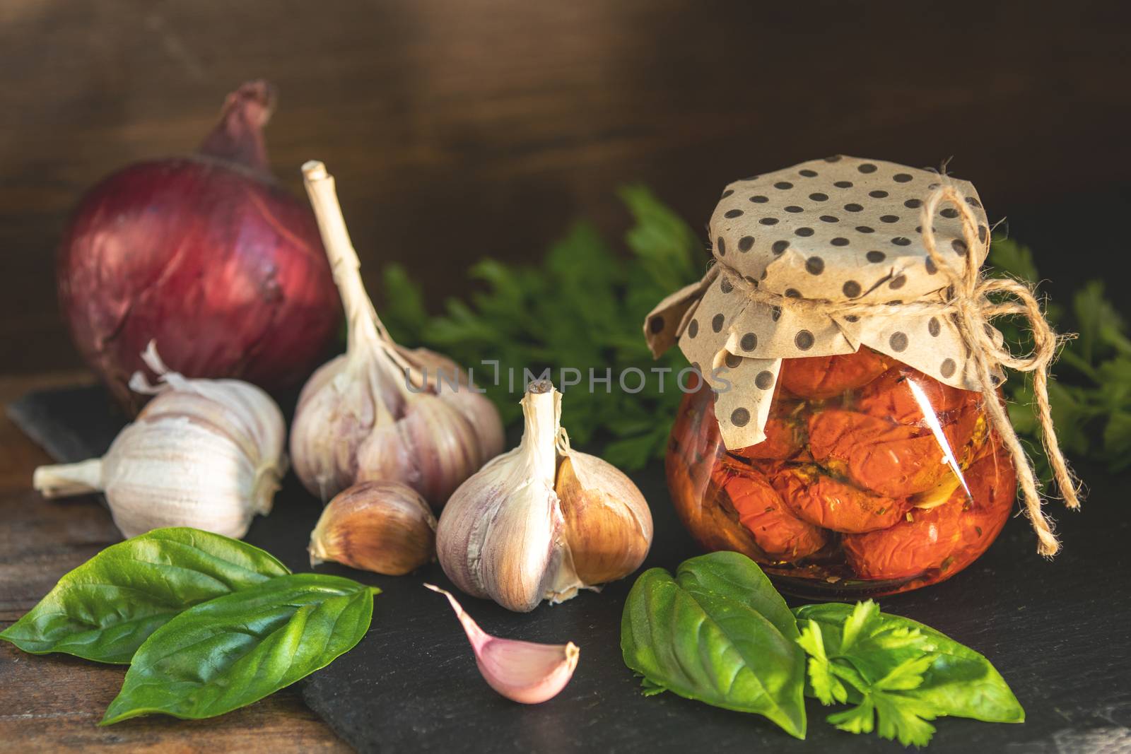 Sun dried tomatoes in glass jar, onion, basil leaves and garlic on cutting board, on wooden background