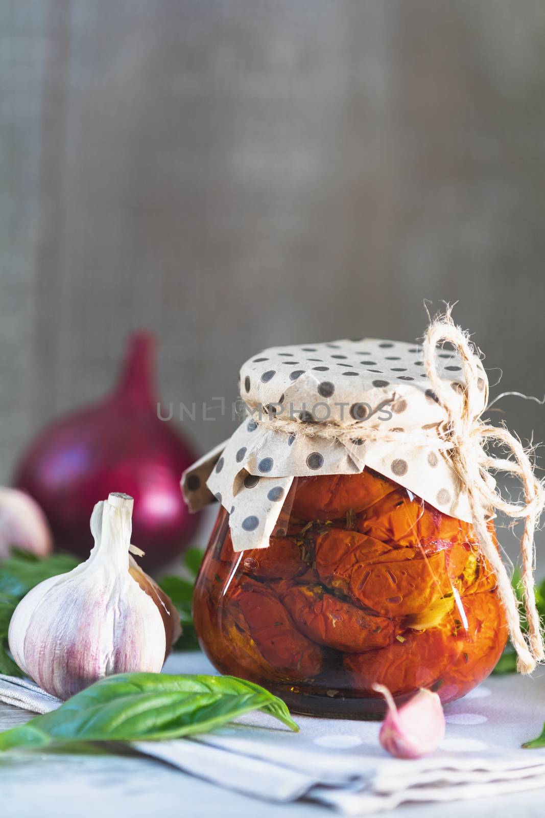 Sun dried tomatoes in glass jar, onion, basil leaves and garlic on cutting board, on wooden background