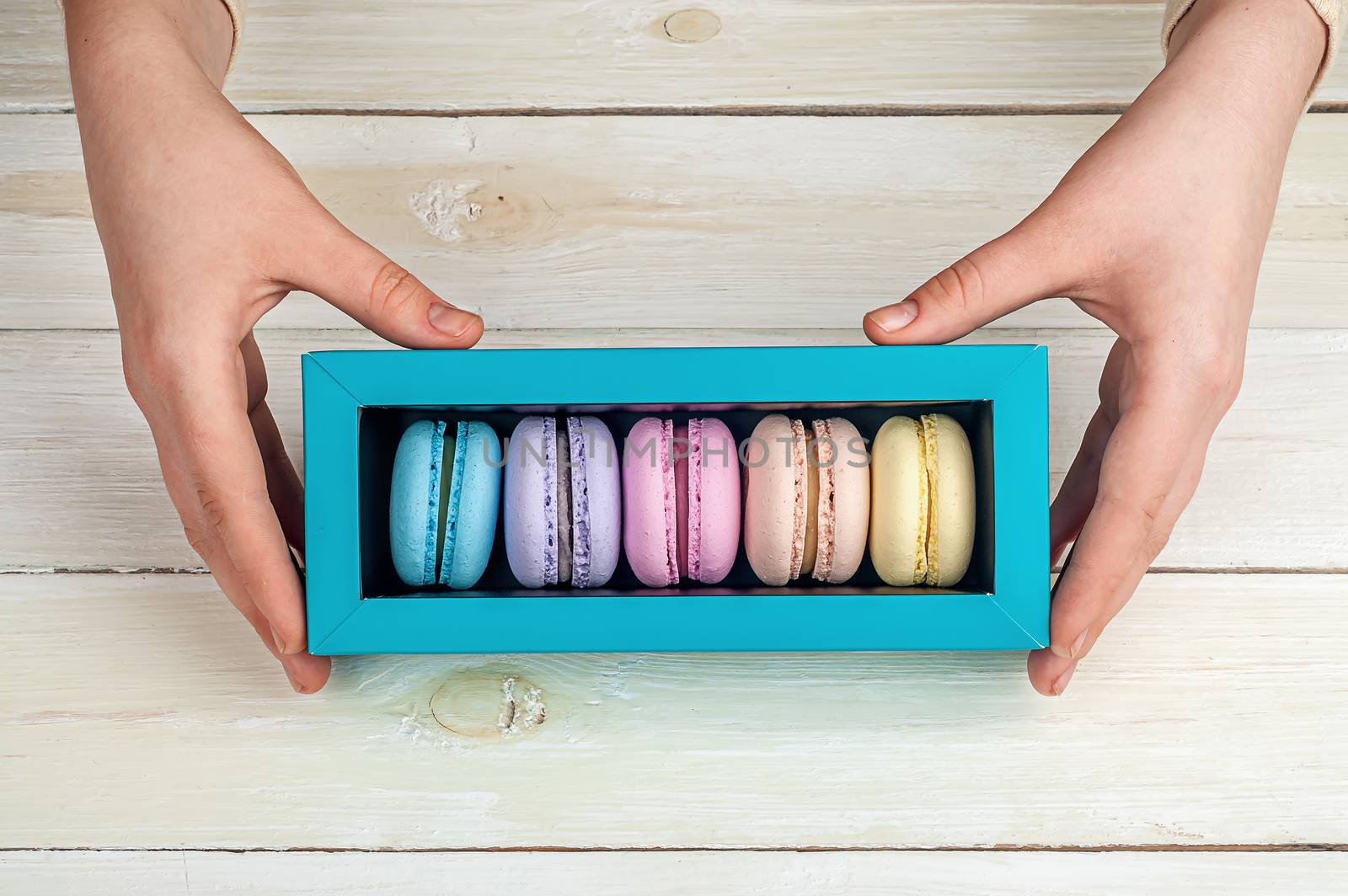 Girl's hands holding box of macaroons on wooden background