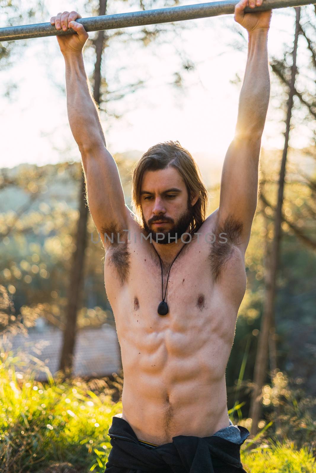 Young bearded man doing pull ups in the park with beautiful sunset on the mountain