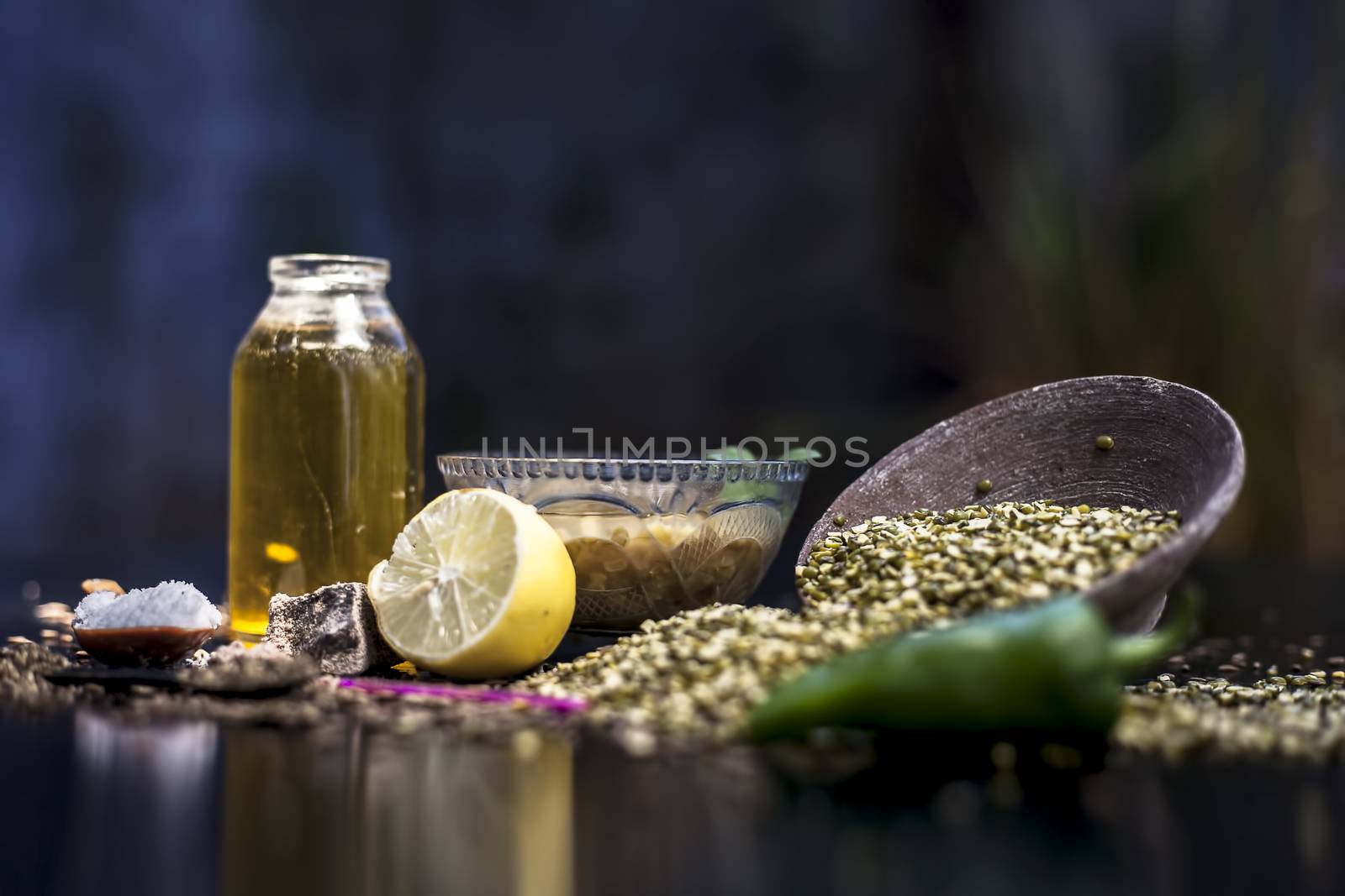 Close up of mung dal soup in glass bowl along with entire constituents with it which are mung bean,green gram, asafoetida, cumin, green chili, black peppercorns, lemon juice and salt on black surface.