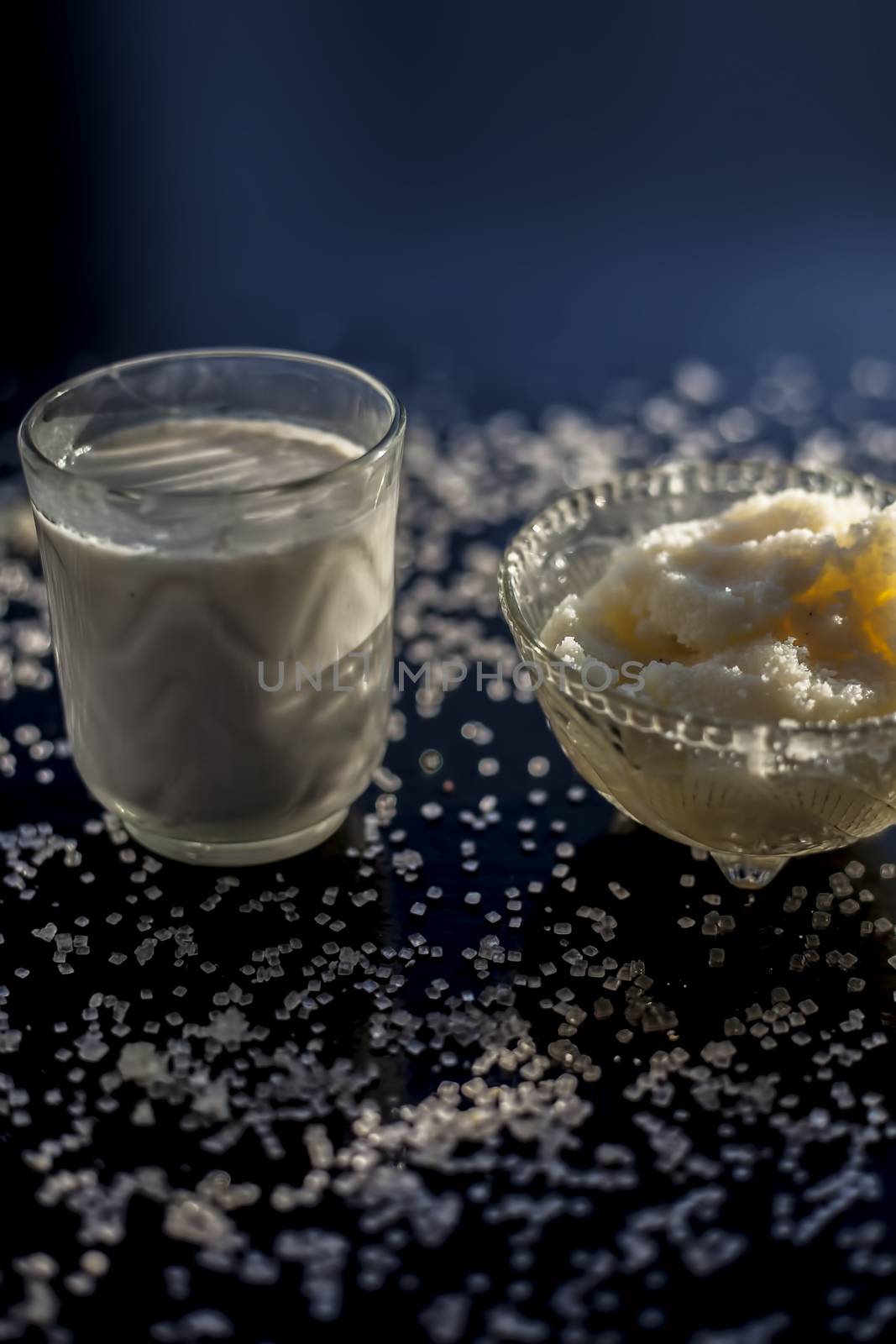 Close up of glass bowl of pure milk well mixed with hot milk in it on black wooden glossy surface along with raw ghee clarified butter and some sugar crystals spread on the surface. Vertical shot. by mirzamlk