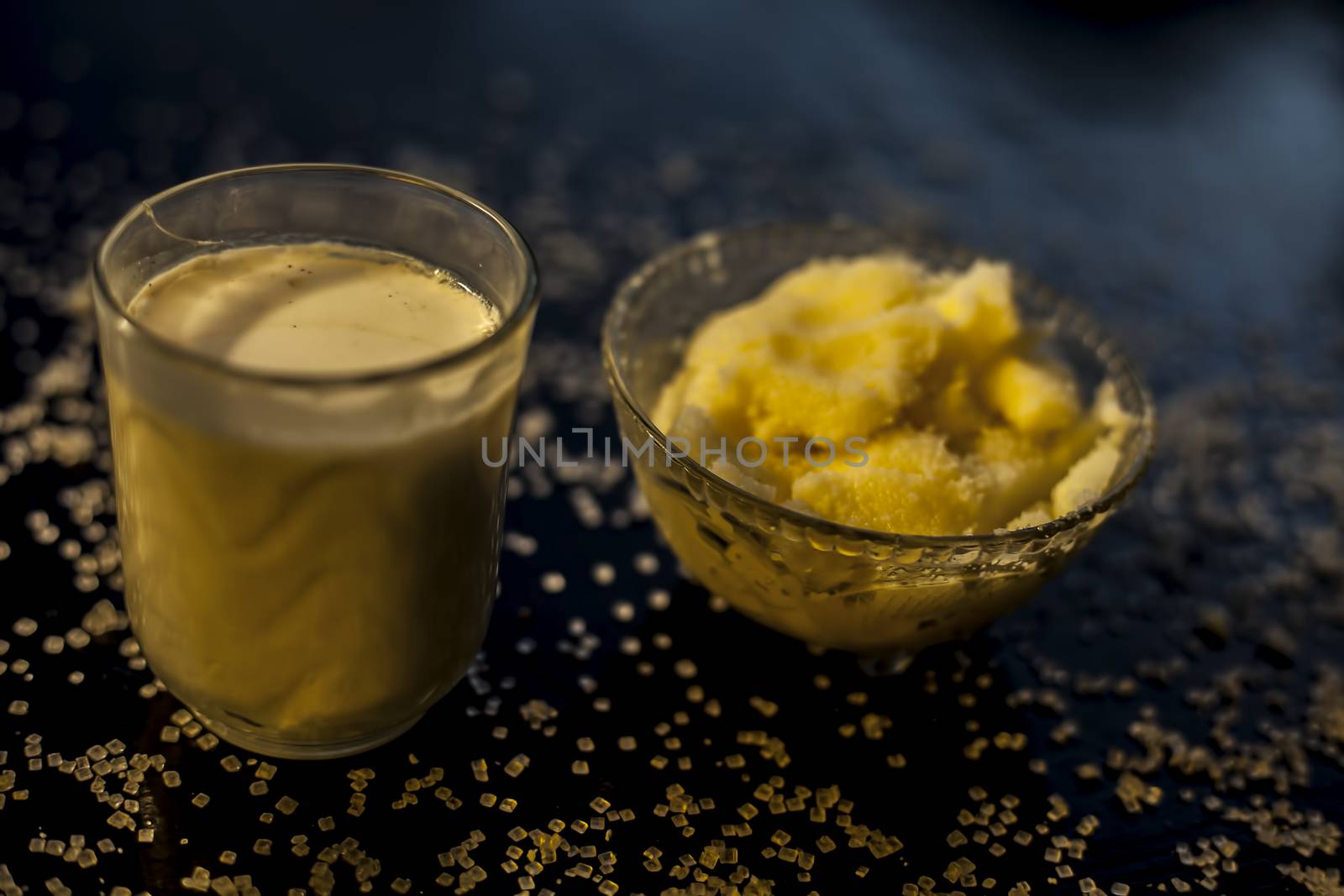 Close up of glass bowl of pure milk well mixed with hot milk in it on black wooden glossy surface along with raw ghee clarified butter and some sugar crystals spread on the surface. Horizontal shot.;