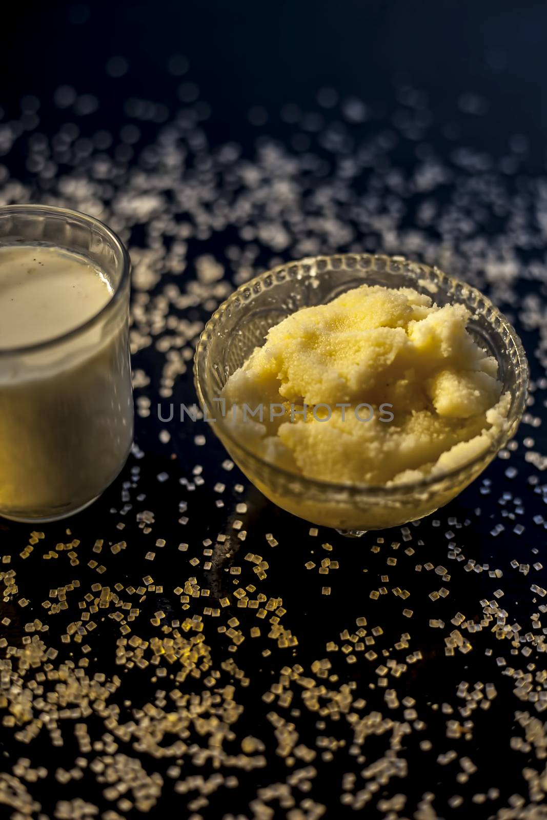Close up of glass bowl of pure milk well mixed with hot milk in it on black wooden glossy surface along with raw ghee clarified butter and some sugar crystals spread on the surface. Vertical shot.