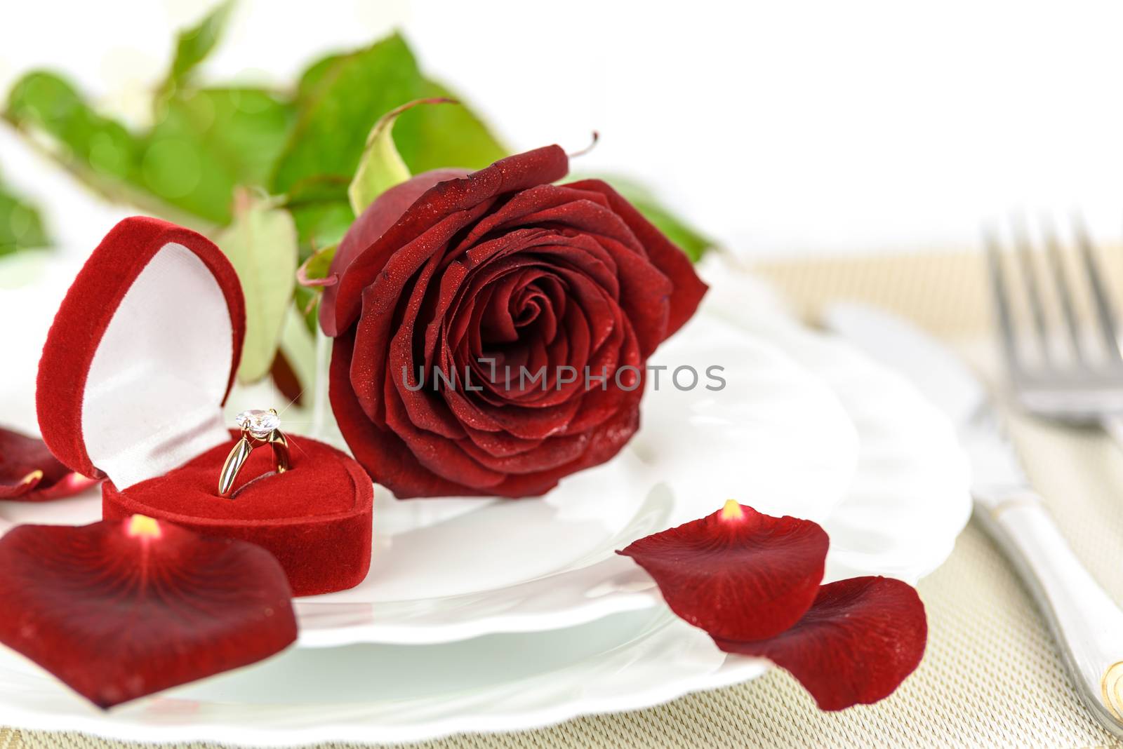 Table setting for a romantic dinner  with Gold engagement ring with diamonds in an elegant velvet box and a red rose.