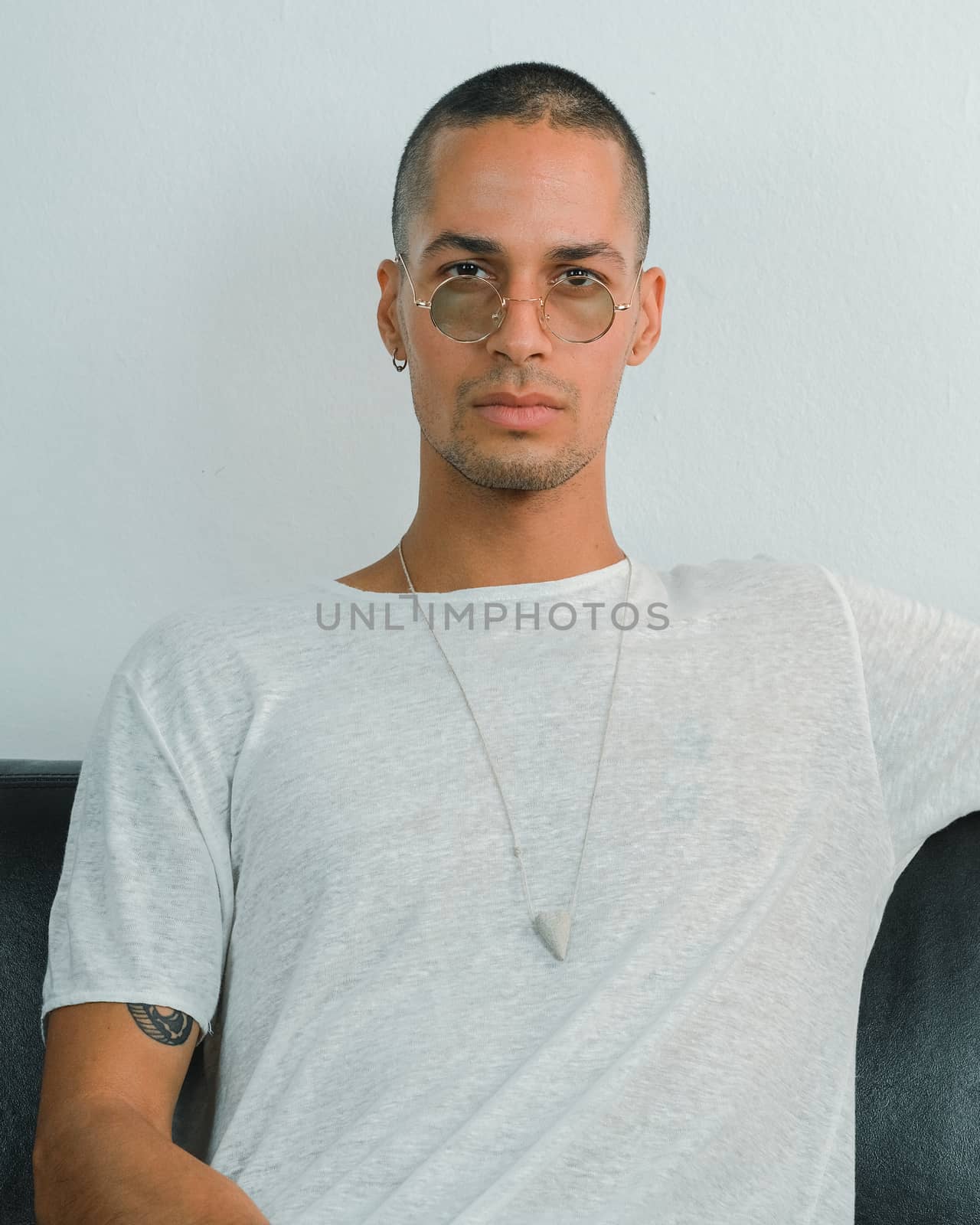 Exotic caucasian young man with round transparent sunglasses looking to the camera while sitting on a sofa with white background