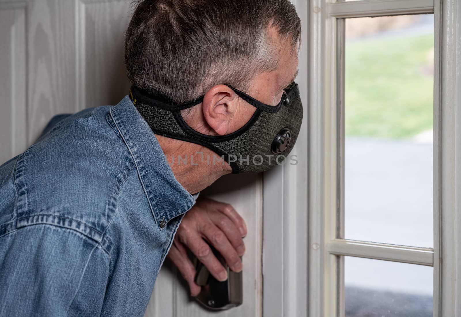 Concerned and worried man wearing a protective breathing mask against flu and coronavirus and looking out of front door