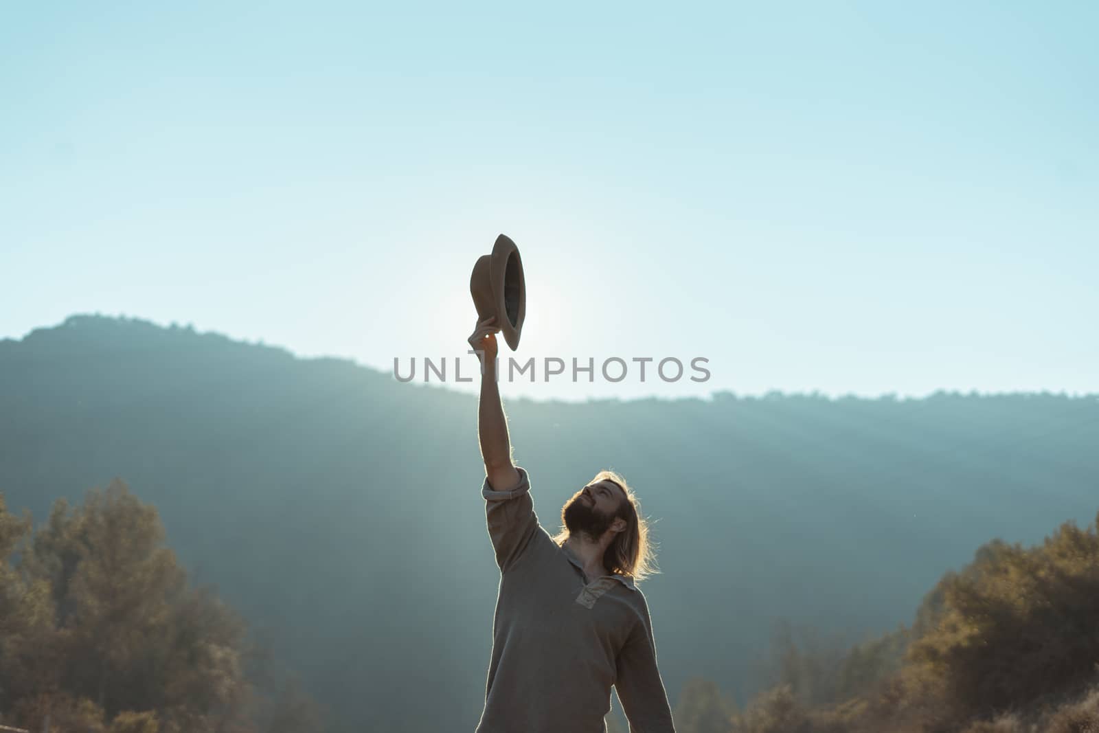 Young man with a beard is holding a hat on a beautiful sunset in the mountains