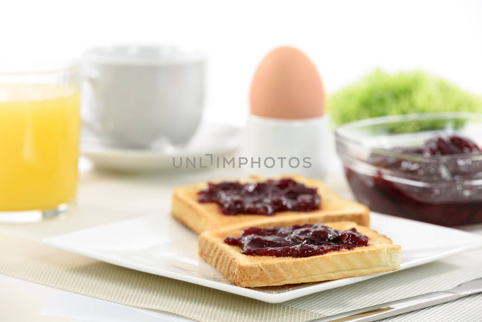 Crispy toast with raspberry jam on a plate and a soft-boiled egg next to orange juice and cup of coffee and apples on white background.