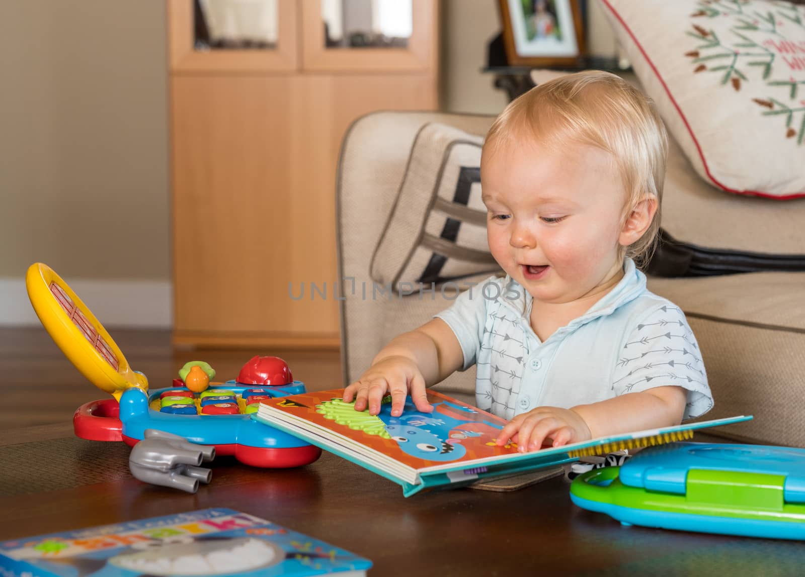 Baby boy sitting on the floor of living room and happily reading a book rather than play with toy computer