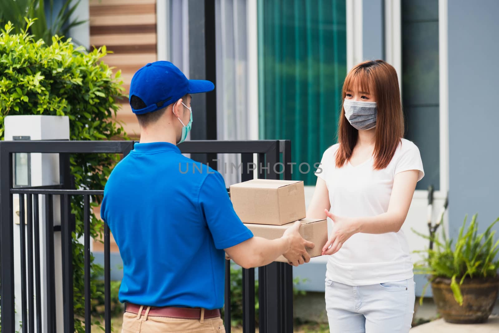 Asian young delivery man courier shopping online give package post box he protective face mask service woman customer receiving box at front home, under curfew quarantine pandemic coronavirus COVID-19