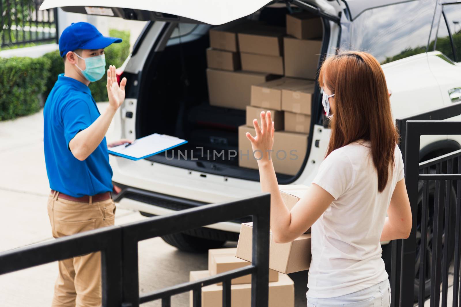 Asian young delivery man raises hand for bye after woman customer received boxes, both protective face mask under curfew quarantine pandemic coronavirus COVID-19 at front home door