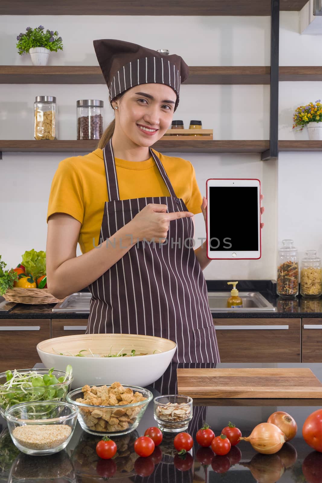The housewife dressed in an apron and a hair cap, holding blank screen tablet computer. Morning atmosphere in a modern kitchen.