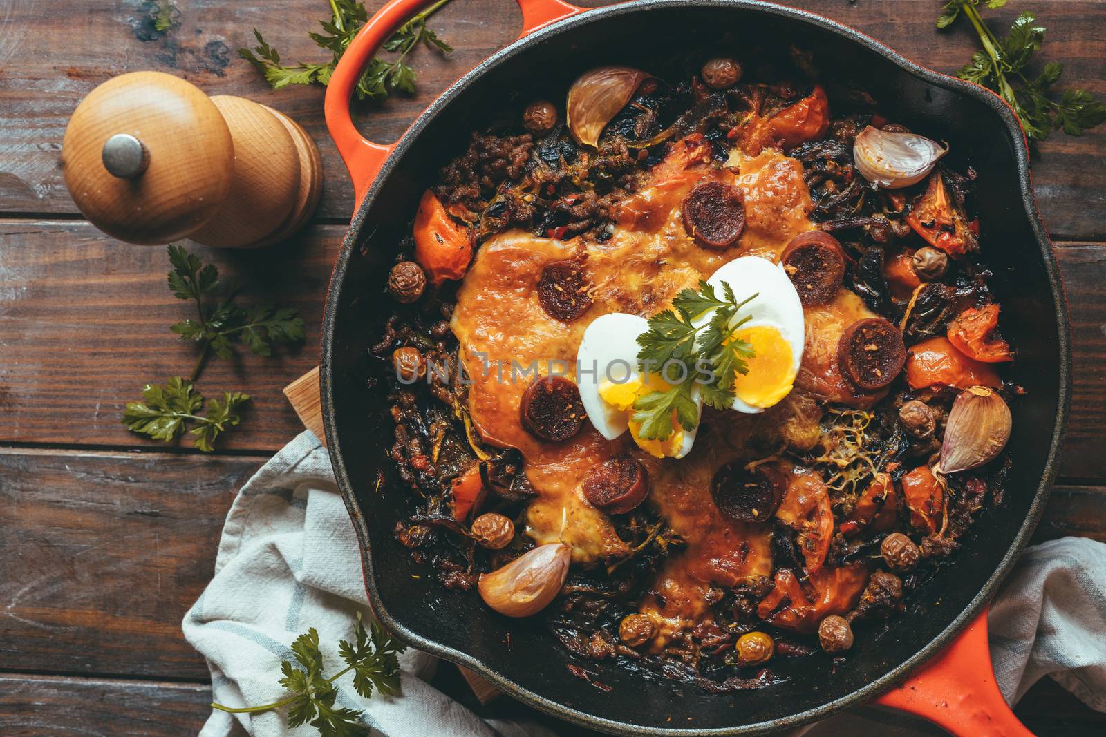 Wrought iron skillet with vegetables, meat, egg, gratin cheese and coriander on a rustic wooden table