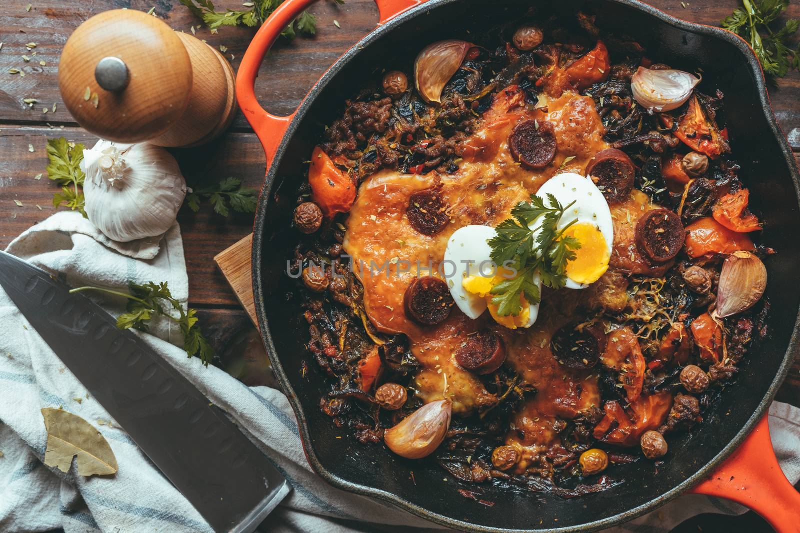 Wrought iron skillet with vegetables, meat, egg, gratin cheese and coriander with a knife on a rustic wooden table