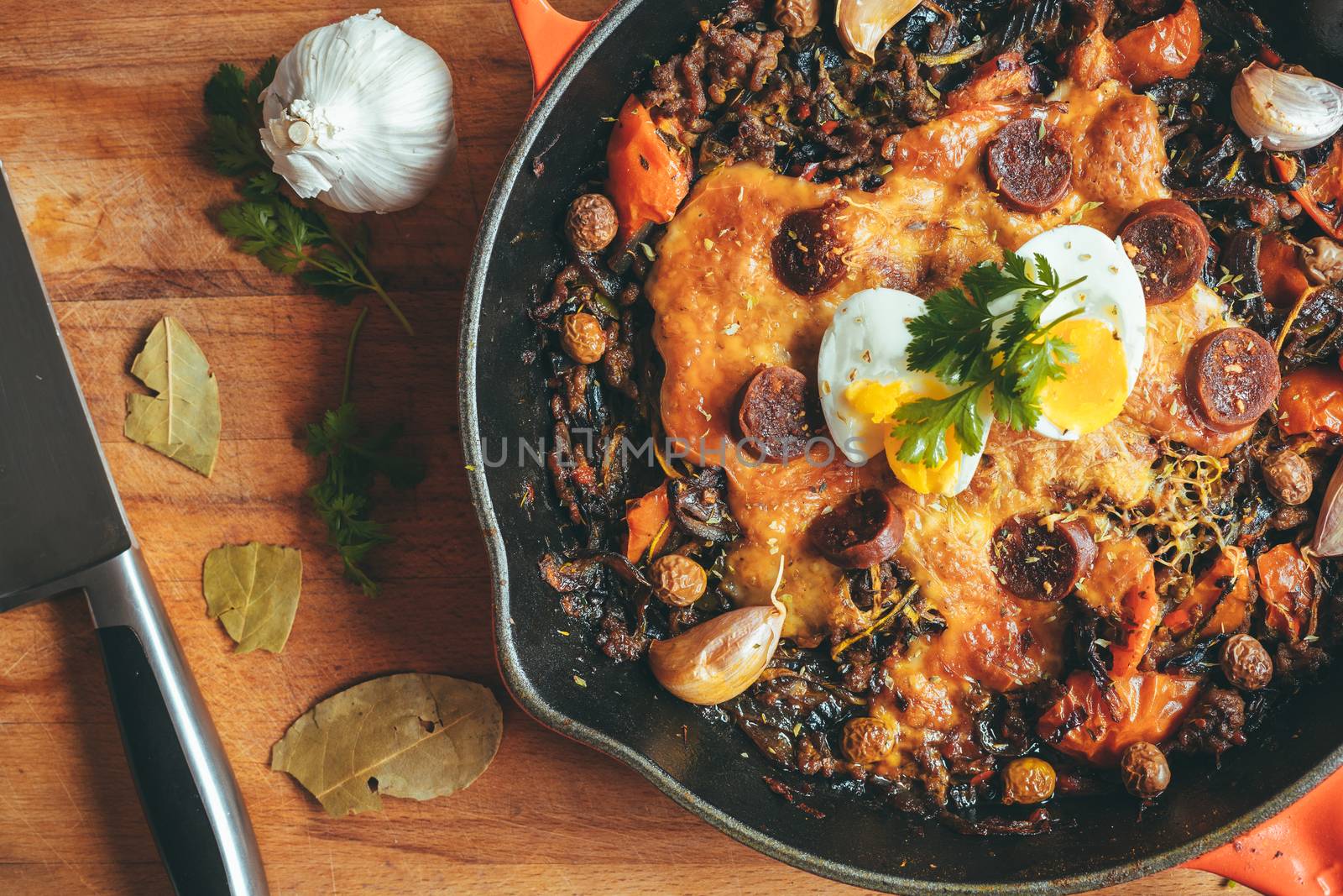 Wrought iron skillet with vegetables, meat, egg, gratin cheese and coriander with a knife on a rustic wooden table