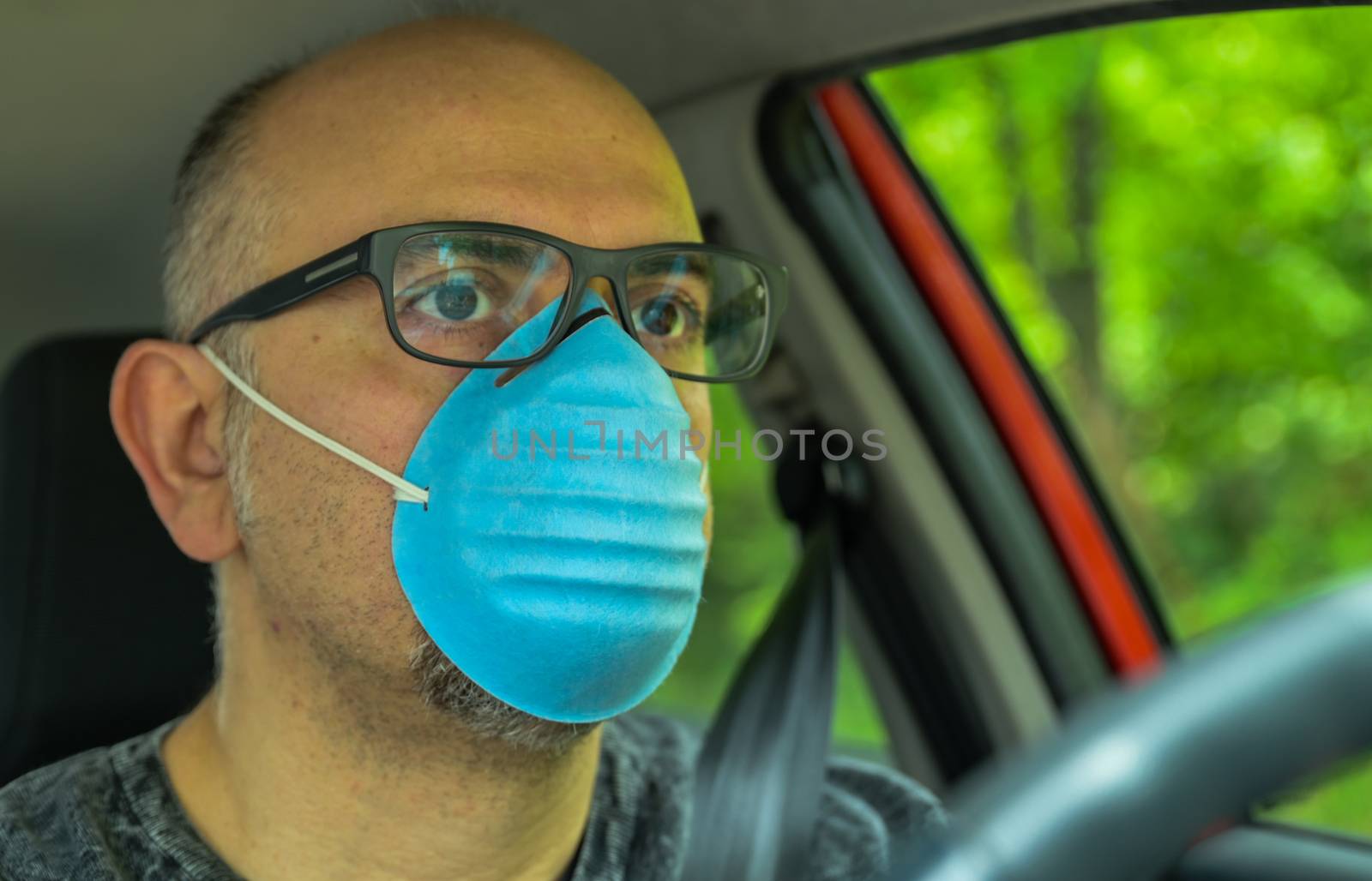 Turin, Piedmont, Italy. April 2020. Coronavirus pandemic: portrait of a Caucasian man driving the car wearing a blue mask to avoid contagion. Selective focus on man and blurred background.