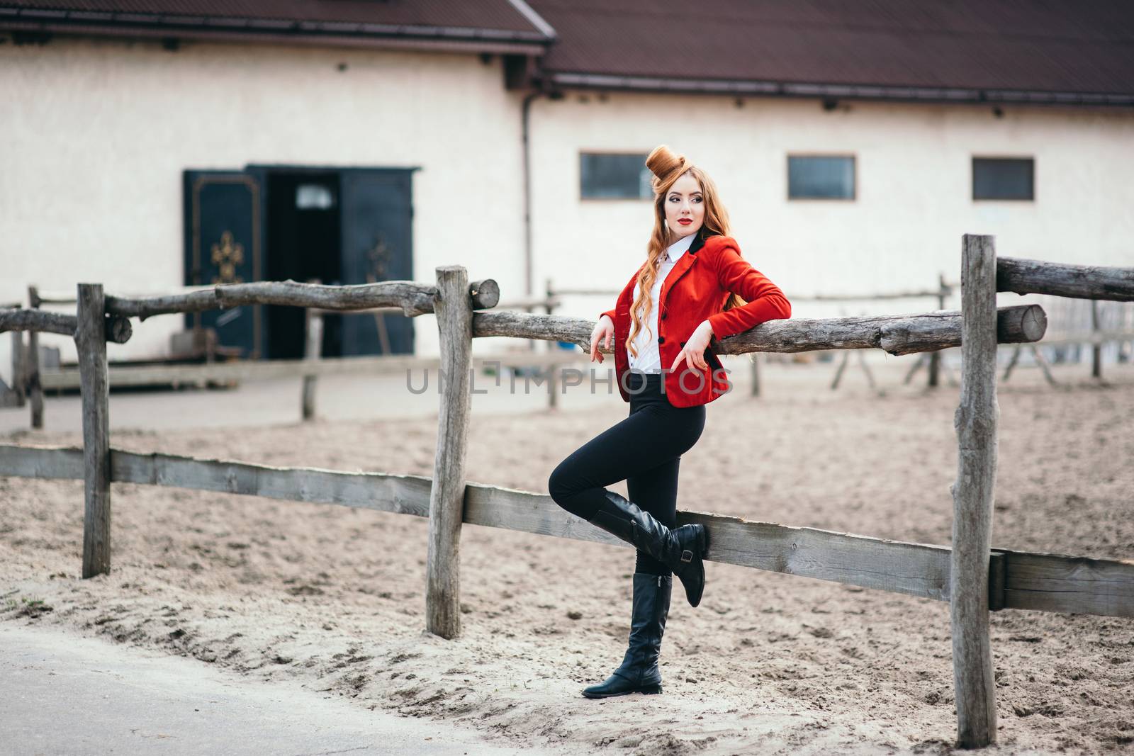 red-haired jockey girl in a red cardigan and black high boots stares at the fence