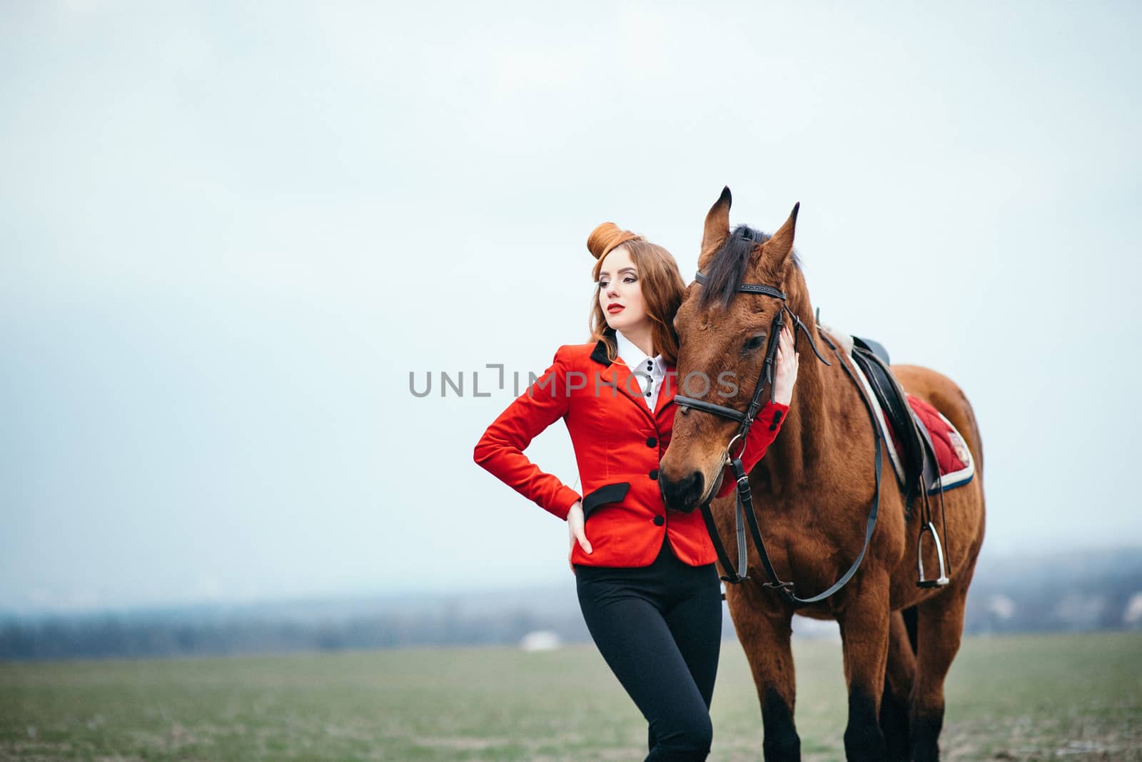 red-haired jockey girl in a red cardigan and black high boots with a horse for a walk