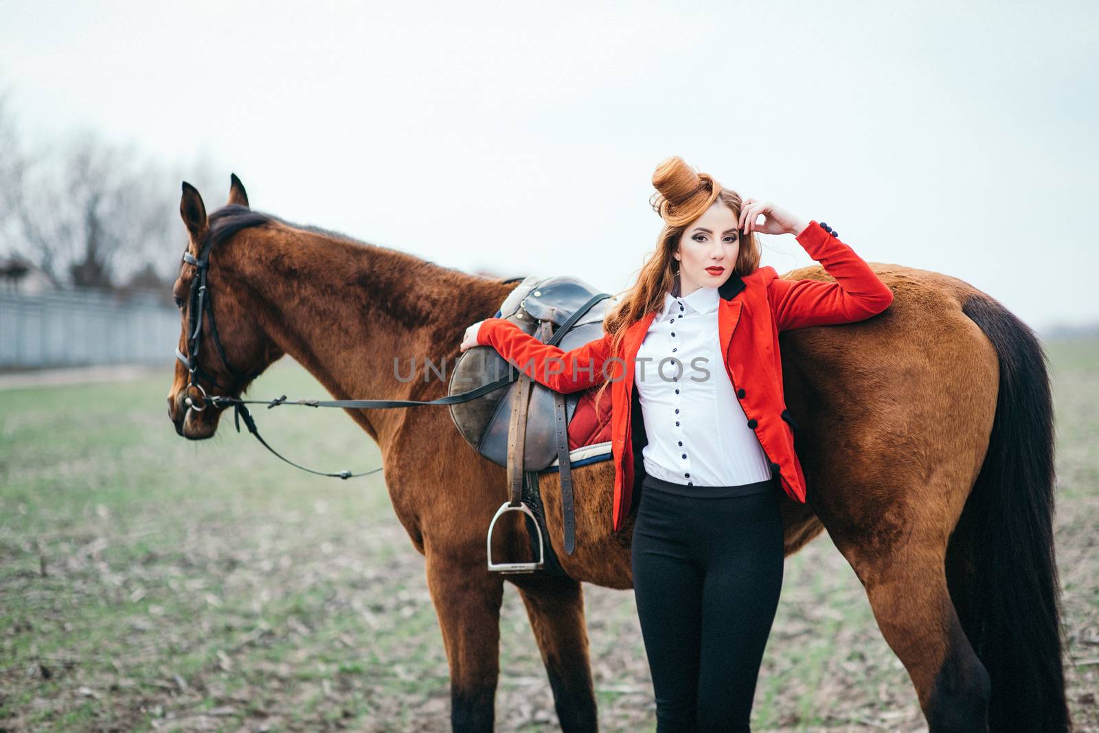 red-haired jockey girl in a red cardigan and black high boots with a horse for a walk