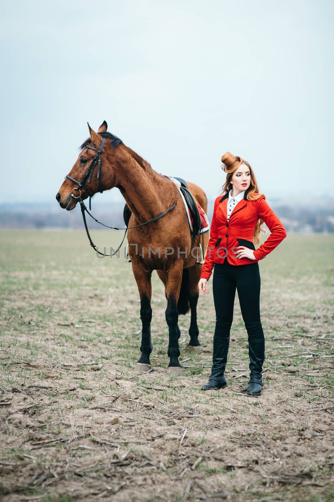 red-haired jockey girl in a red cardigan and black high boots with a horse for a walk