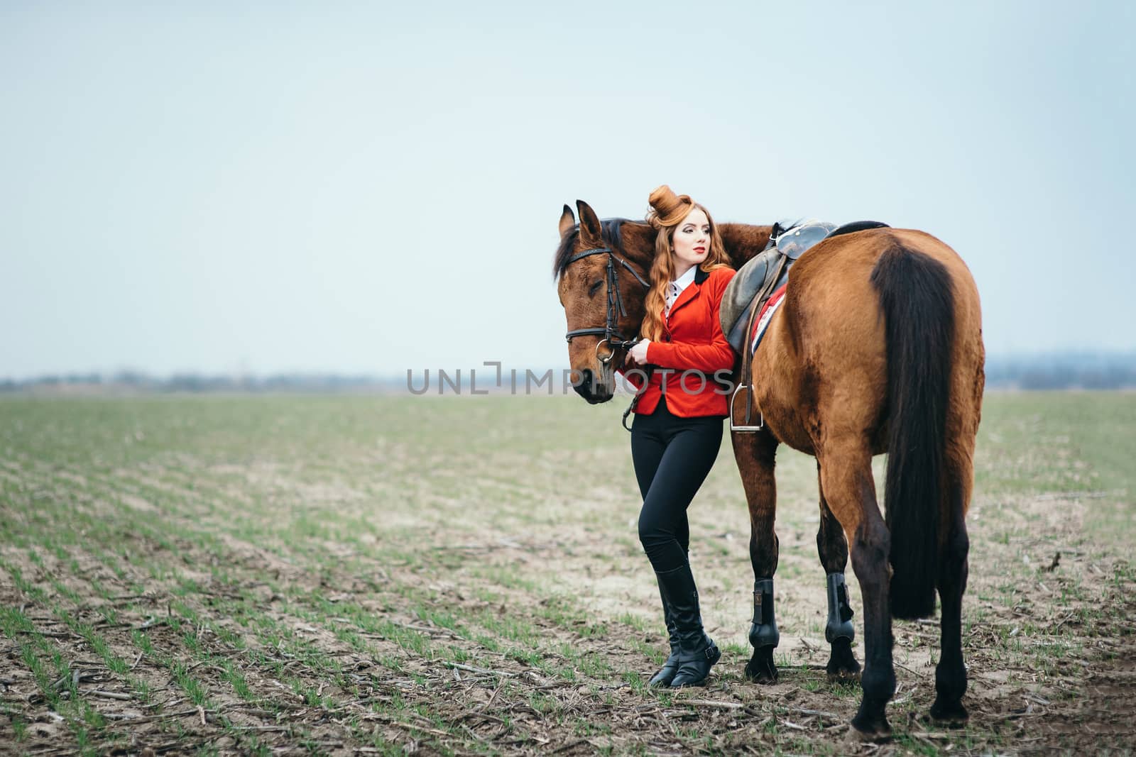 red-haired jockey girl in a red cardigan and black high boots with a horse for a walk