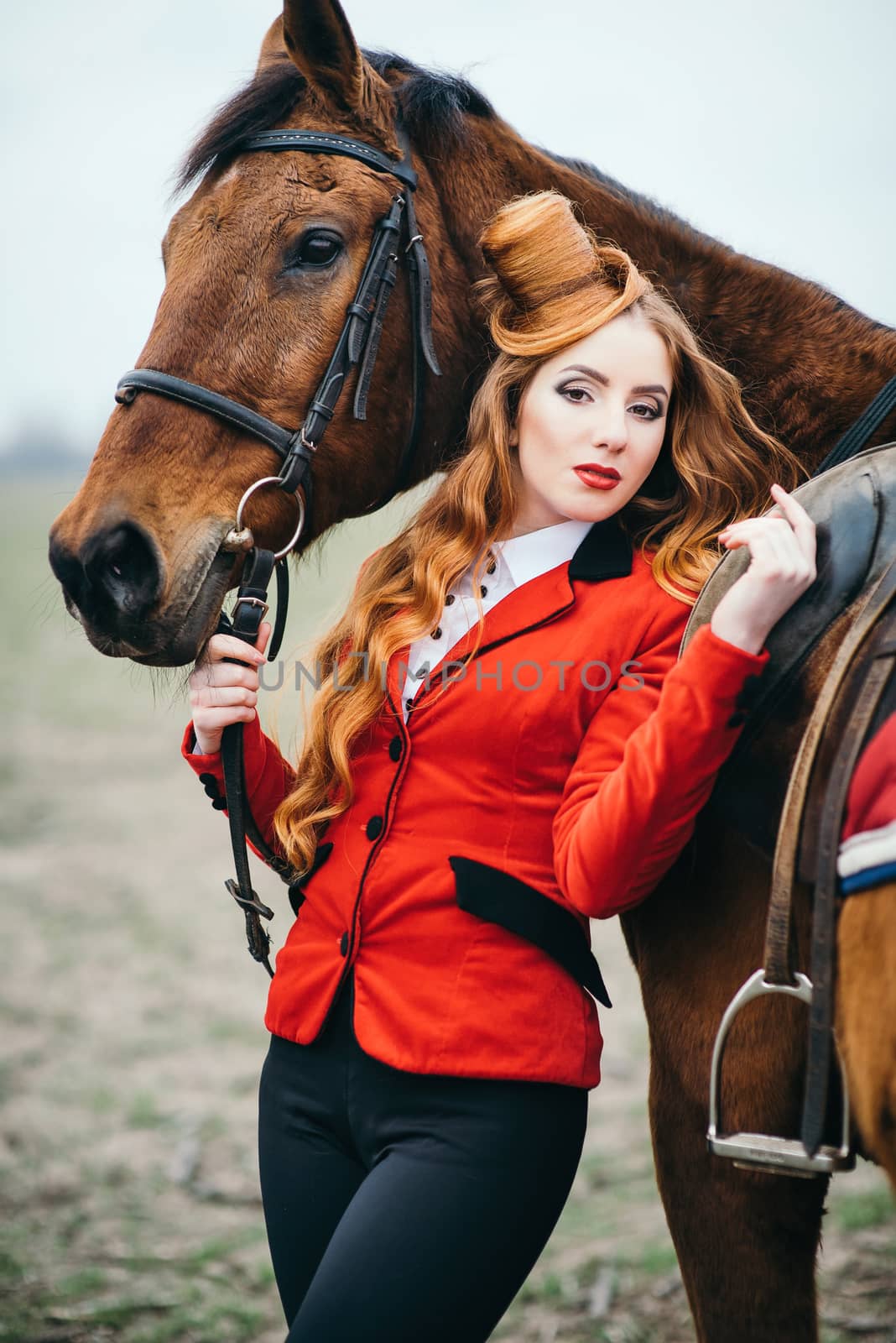 red-haired jockey girl in a red cardigan and black high boots with a horse for a walk