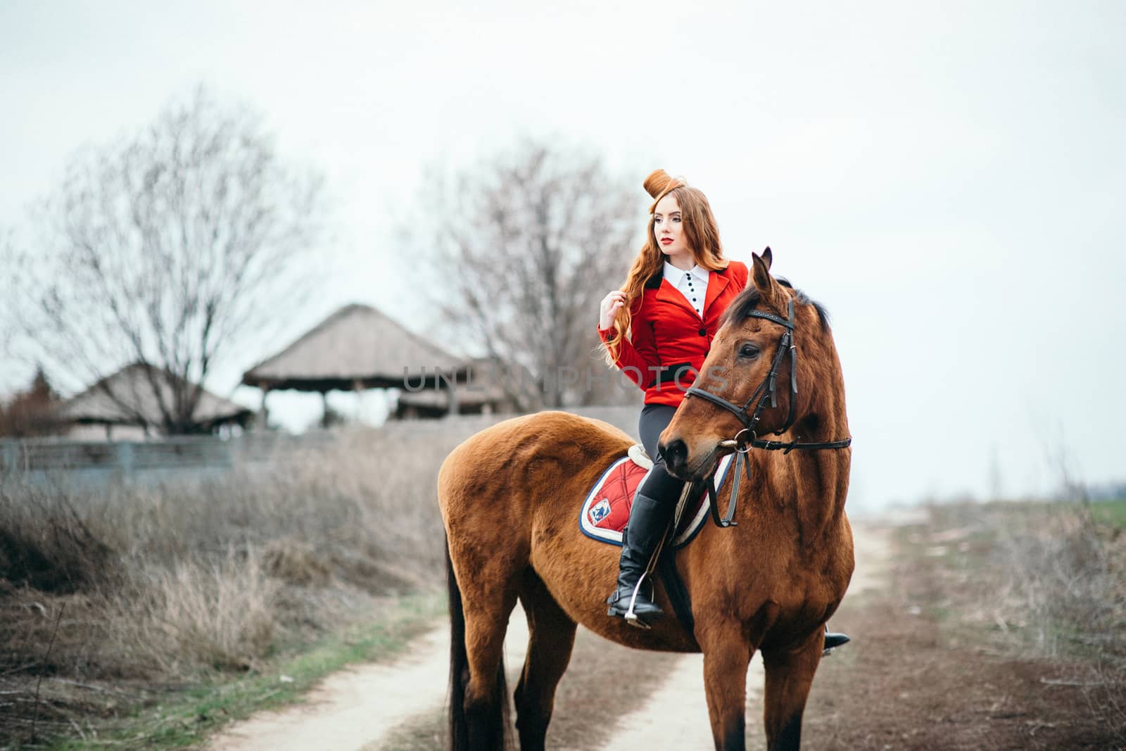 red-haired jockey girl in a red cardigan and black high boots with a horse for a walk