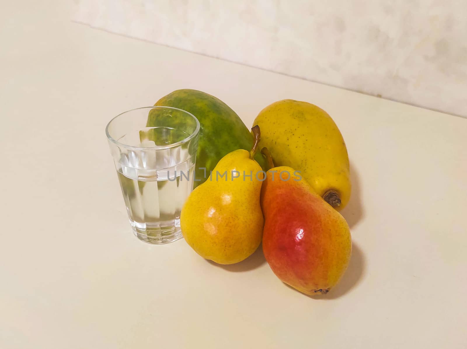 Glass of water and yellow fruits, papaya and pear on white bench in soft light.