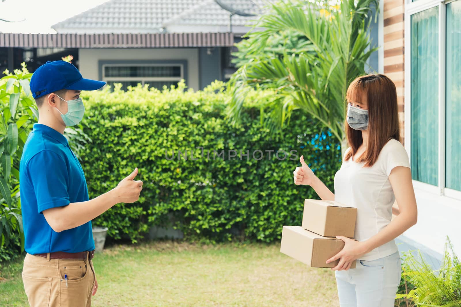 Asian delivery express courier young man giving parcel boxes to woman customer receiving both protective face mask and show thumbs up finger for good support sign, under curfew pandemic coronavirus