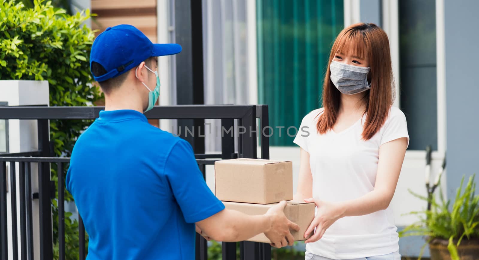 Asian young delivery man courier shopping online give package post box he protective face mask service woman customer receiving box at front home, under curfew quarantine pandemic coronavirus COVID-19