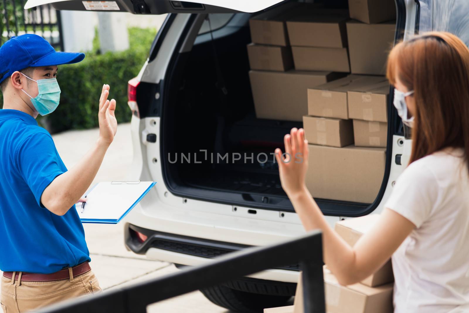 Asian young delivery man raises hand for bye after woman customer received boxes, both protective face mask under curfew quarantine pandemic coronavirus COVID-19 at front home door