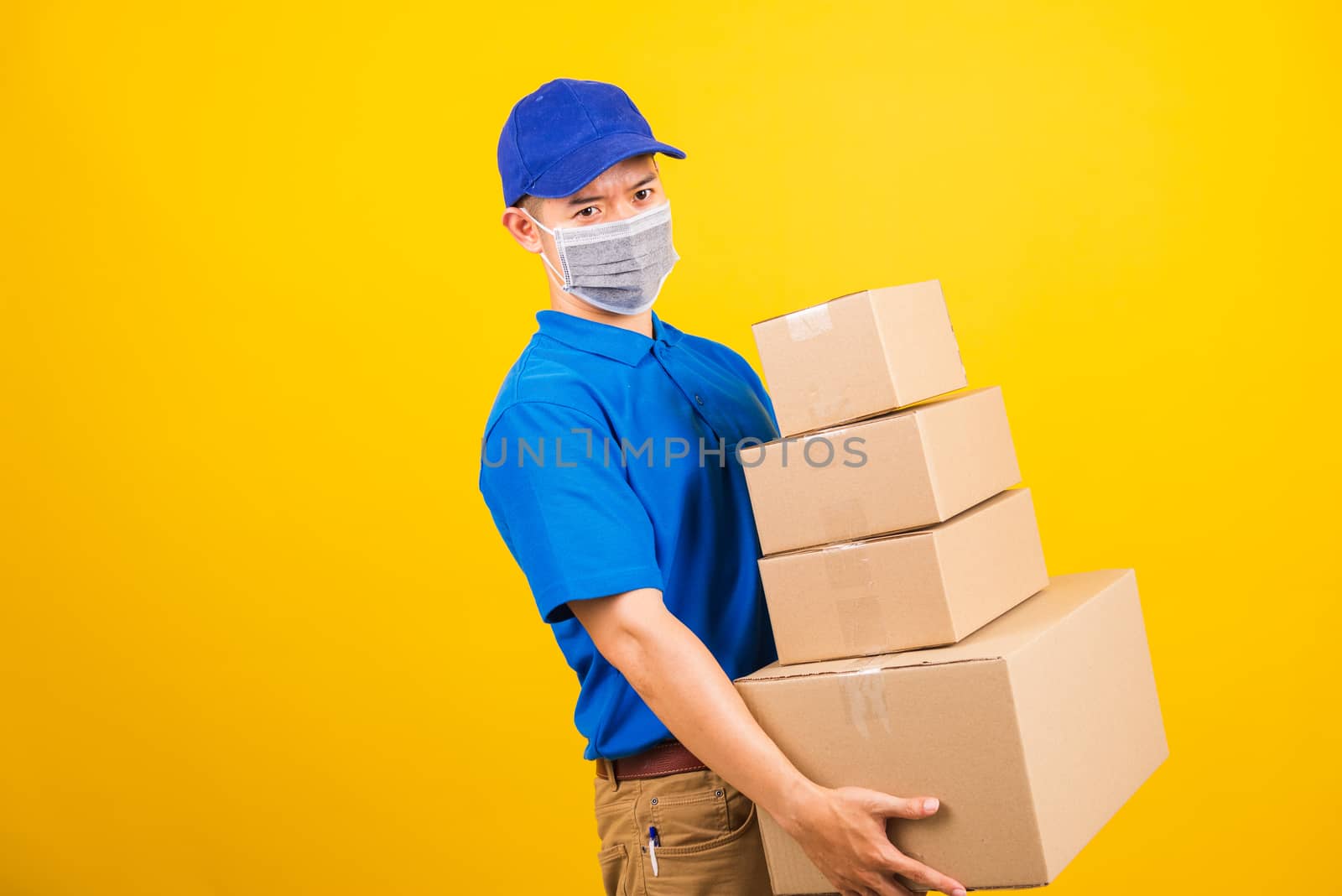 Asian young delivery worker man in blue t-shirt and cap uniform wearing face mask protective lifting stack a lot of boxes, under coronavirus or COVID-19, studio shot isolated yellow background