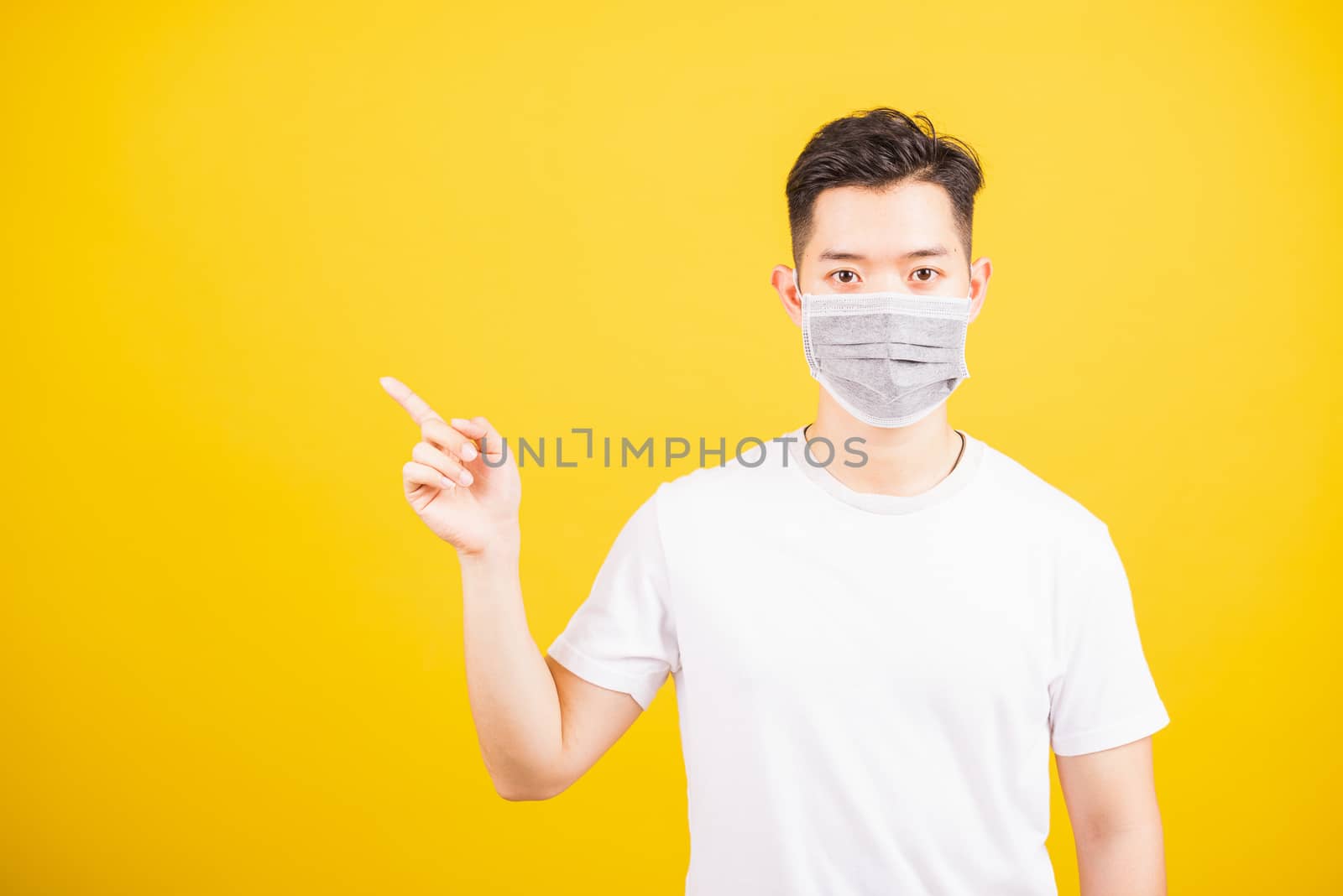 Portrait Asian young man wearing face mask protects filter dust pm2.5 anti-pollution, anti-smog, air pollution and COVID virus her pointing out side on yellow background, with copy space