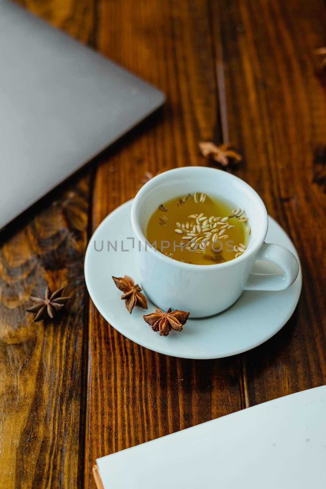 White cup of tea with star anise on a wooden table with a computer in the background