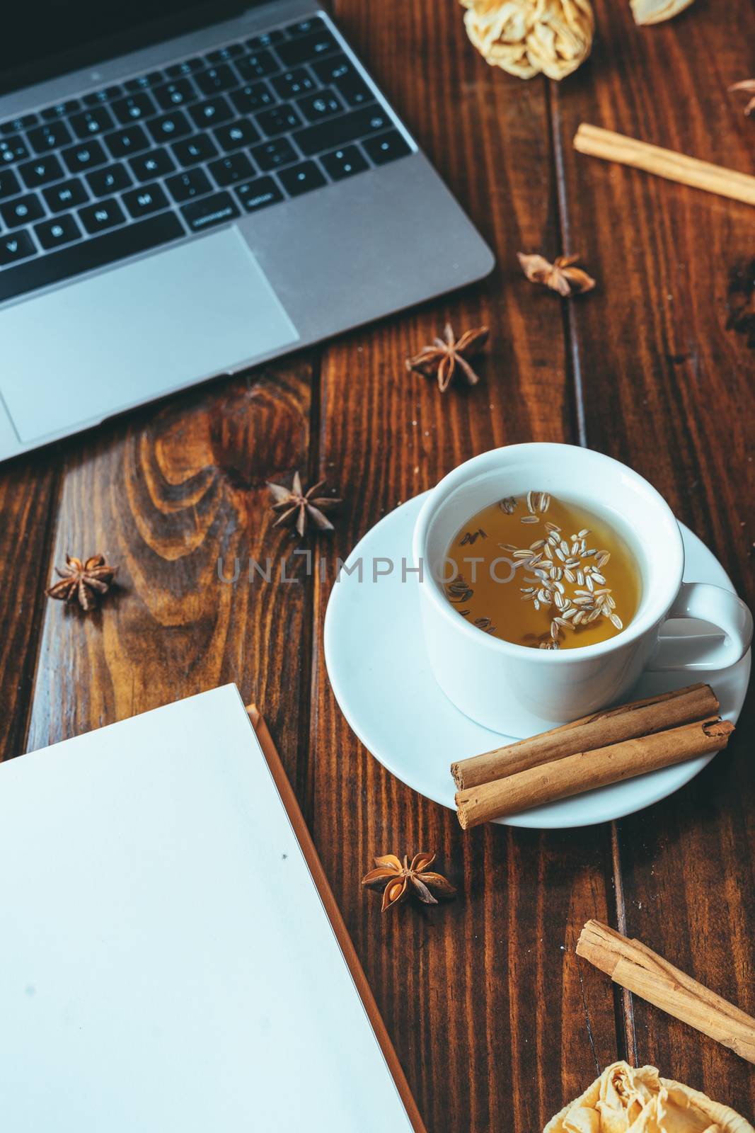 White cup of tea with cinnamon with a notebook on a wooden table with a computer in the background