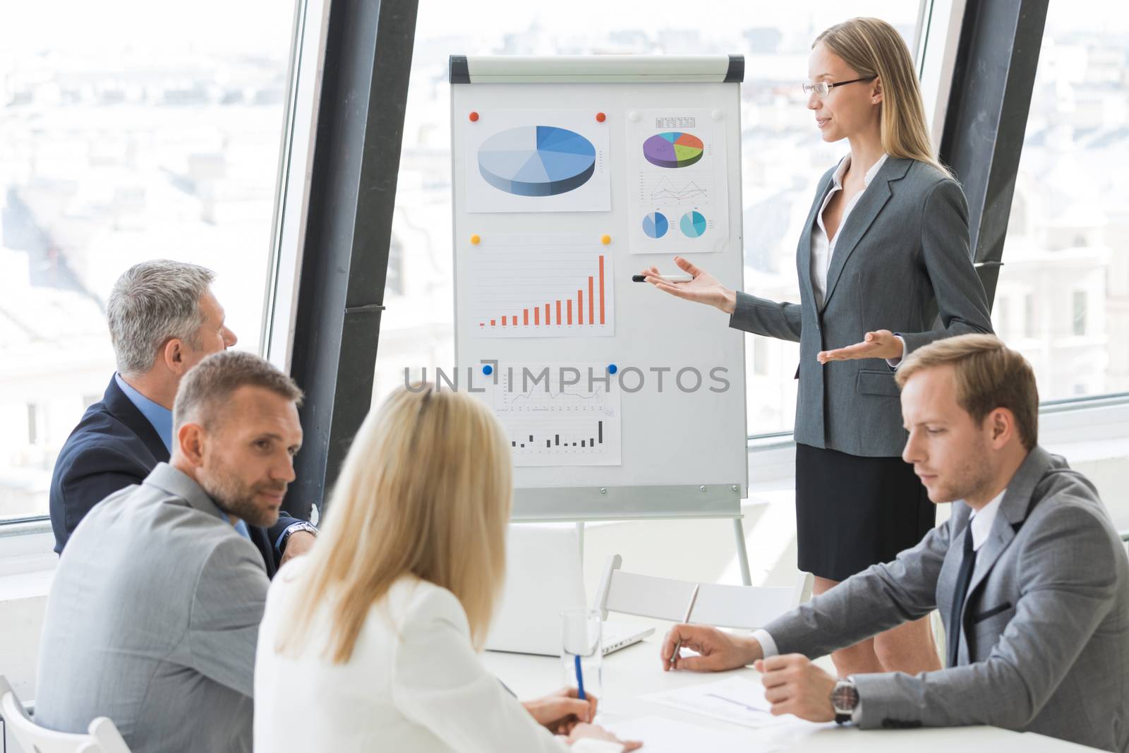 Businesswoman giving presentation using financial diagrams at flipchart to colleagues in office at meeting