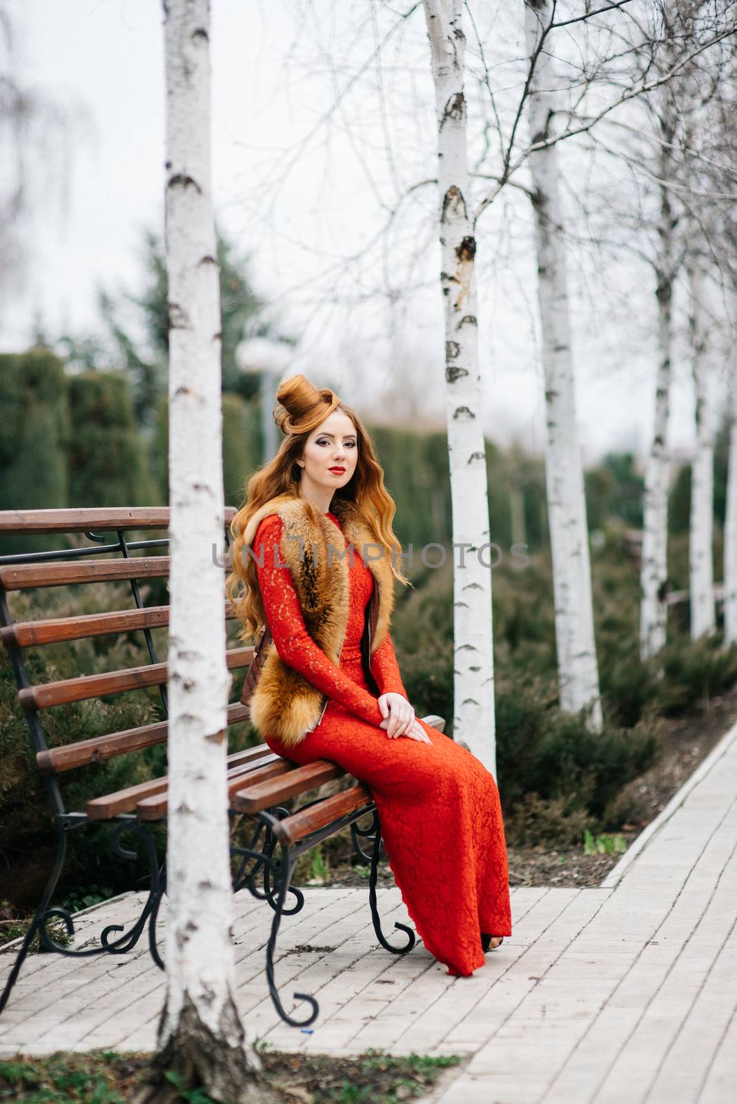 young girl with red hair in a bright red dress on a bench in an empty park among the birches