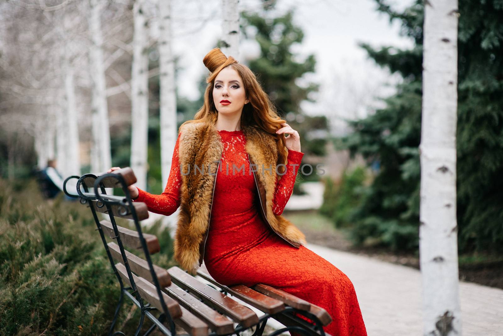 young girl with red hair in a bright red dress on a bench in an empty park among the birches