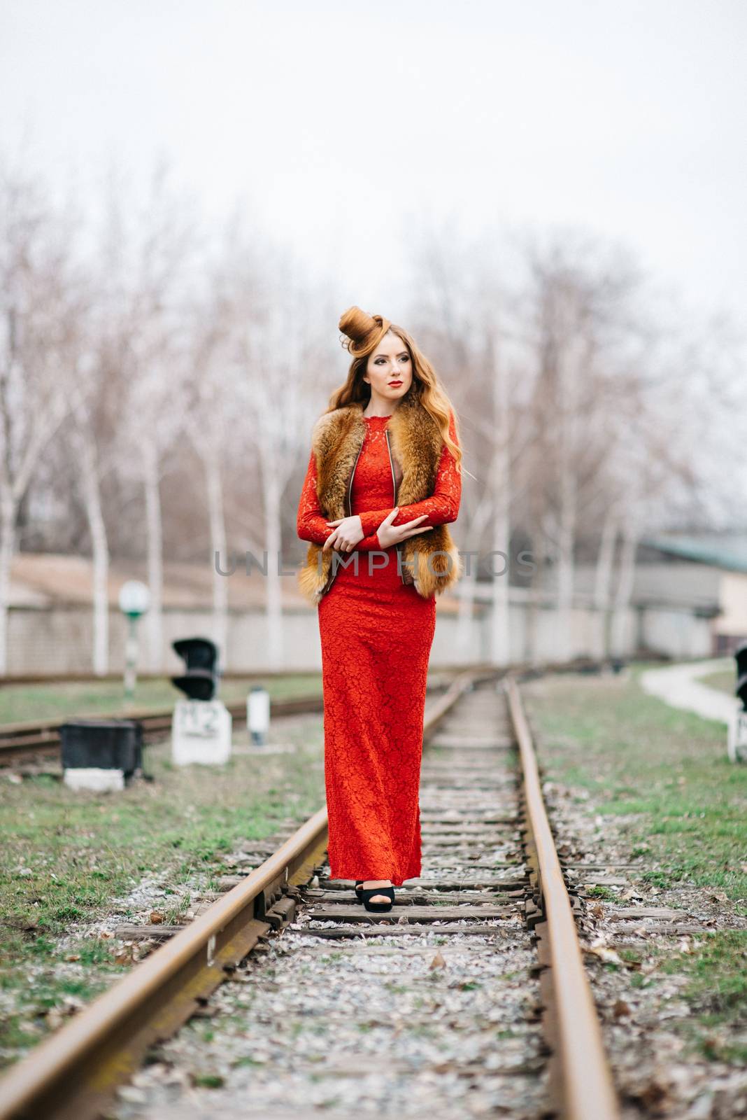 young girl with red hair in a bright red dress on the railway tracks of the station