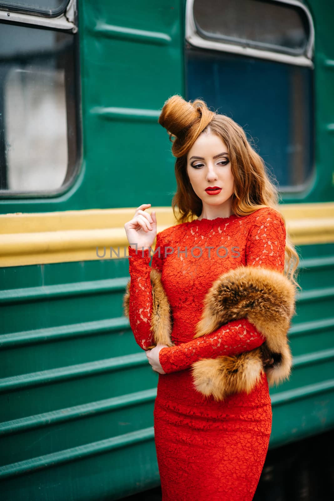 young girl with red hair in a bright red dress near an old green passenger car