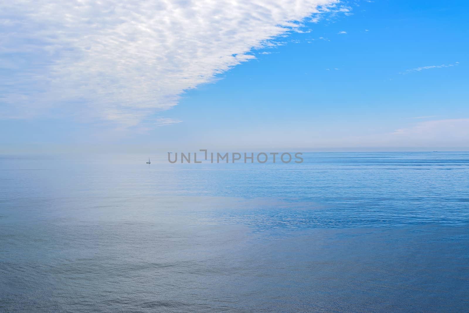 The blue sky and clouds reflect on the calm surface of the ocean and the ships in the distance.