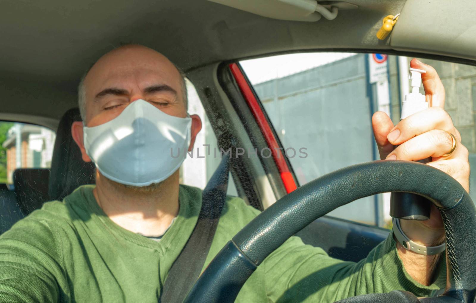 Turin, Piedmont, Italy. April 2020. Caucasian man driving the car wearing a mask. Spray the alcohol to disinfect the surfaces: the drops shine in the air. Accidentally they go into his eyes.